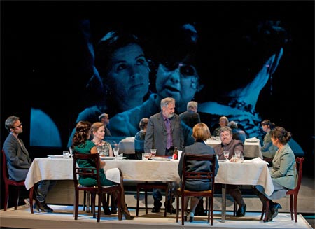 The dinner table of THE PATRIARCH. The big screen reveals rifts in the amiable atmosphere: daughter-in-law (Kristiina Halttu, left) and wife (Kirsti Wallasvaara). Photo courtesy: Stefan Bremer